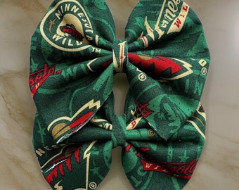 Pode incluir: Um laço de tecido verde e vermelho com o logotipo do Minnesota Wild repetido no tecido. O logotipo inclui as palavras "Minnesota Wild" e uma imagem estilizada de um animal selvagem.