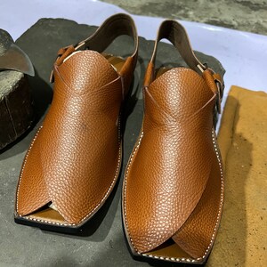May include: A pair of brown leather sandals with a textured surface. The sandals have a unique design with a strap around the ankle and a buckle closure. The edges are stitched with white thread. The sandals are displayed on a dark gray surface.