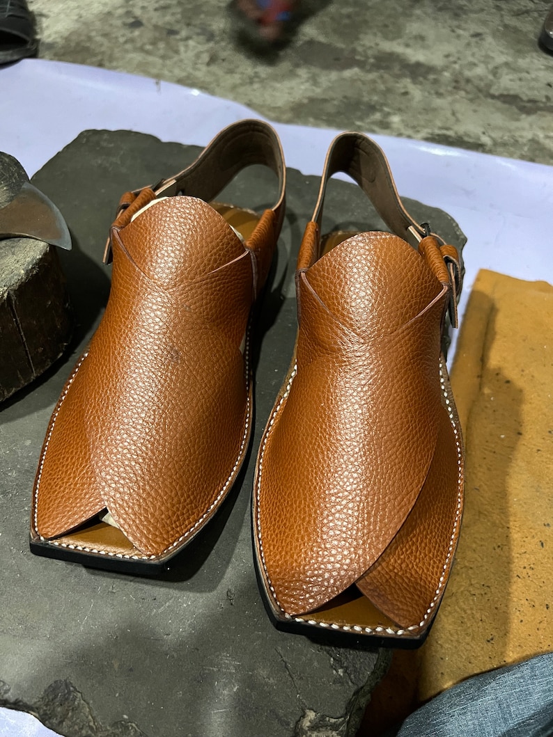 May include: A pair of brown leather sandals with a textured surface. The sandals have a unique design with a strap around the ankle and a buckle closure. The edges are stitched with white thread. The sandals are displayed on a dark gray surface.