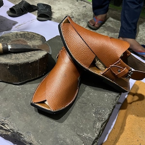 May include: A pair of brown leather sandals with a unique design, featuring a strap and buckle closure. The sandals are displayed on a rough, gray stone surface, showcasing the craftsmanship and style of the footwear.