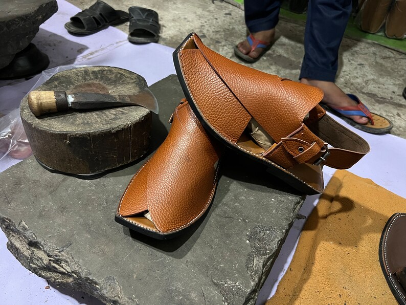 May include: A pair of brown leather sandals with a unique design, featuring a strap and buckle closure. The sandals are displayed on a rough, gray stone surface, showcasing the craftsmanship and style of the footwear.