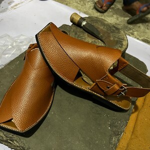 May include: A pair of brown leather sandals with a textured finish. The sandals have a square toe design and a strap with a buckle closure. White stitching accents the edges. The sandals are displayed on a dark gray surface.