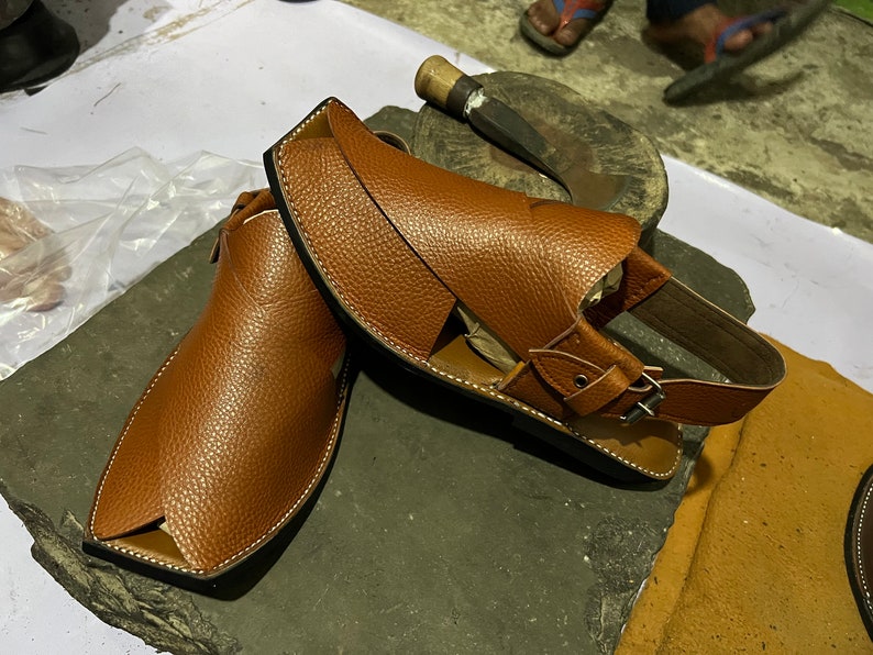May include: A pair of brown leather sandals with a textured finish. The sandals have a square toe design and a strap with a buckle closure. White stitching accents the edges. The sandals are displayed on a dark gray surface.