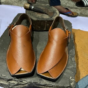 May include: A pair of brown leather Peshawari sandals with a textured finish. The sandals feature a strap around the ankle and a cross-over design on the top. The soles are black with white stitching. The sandals are displayed on a dark gray stone surface.