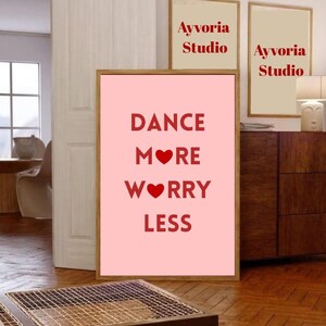 Peut inclure: Une affiche rose encadrée avec les mots "DANCE MORE WORRY LESS" en rouge. Deux petites affiches encadrées avec le texte "Ayyvoria Studio" sont également visibles. L'œuvre est exposée dans une pièce avec des planchers et des meubles en bois.