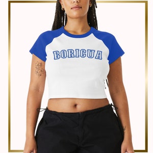 Puede incluir: Camiseta blanca y azul recortada con un dise&ntilde;o de estilo b&eacute;isbol. La camiseta tiene un dise&ntilde;o de bloque de color azul y blanco y dice "BORICUA" en letras azules.