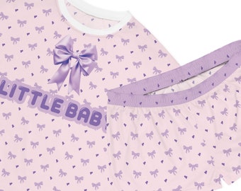 Könnte beinhalten: Rosa-lilafarbener Pyjama-Set mit Schleife und dem Text "LITTLE BAB" auf dem Oberteil. Das Set hat ein Muster aus kleinen Herzen und Schleifen.
