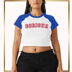 Puede incluir: Una camiseta blanca recortada con mangas ragl&aacute;n azules y la palabra "BORICUA" impresa en rojo en la parte delantera.