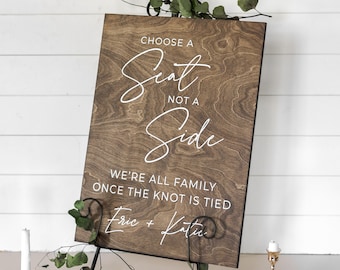 May include: A wooden sign with the text "Choose a Seat, Not a Side. We're all family once the knot is tied. Eric + Katie" in white lettering.