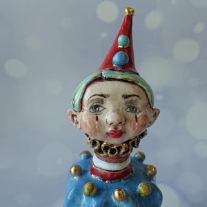 May include: A whimsical ceramic figurine of a clown with a red conical hat adorned with blue and gold accents. The clown has a pale face with red lips, teardrops, and a ruffled collar. The blue outfit is decorated with gold spheres.