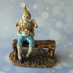 Pode incluir: Uma estatueta de cerâmica de uma figura com asas, usando um chapéu de festa dourado, camisa laranja e jardineiras azuis, sentada num banco de madeira castanho. A figura segura uma pequena chávena de chá branca. A base tem textura de seixos.