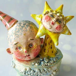 May include: A ceramic sculpture featuring a pale, round-faced figure with a pink and green striped cone hat, positioned next to a bright yellow sun-shaped figure with a human face. The base is decorated with small, light blue spiral accents.
