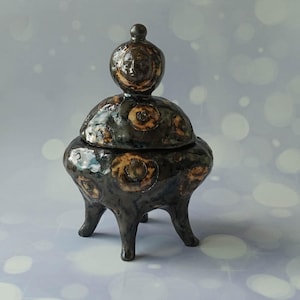 Può includere: Un barattolo in ceramica scuro e lucido con coperchio e tre gambe. La superficie presenta un motivo maculato di cerchi neri e marrone chiaro. Il coperchio è sormontato da un pomello sferico che mostra un volto umano scolpito sul davanti.