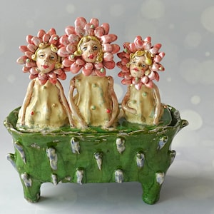 Puede incluir: Una escultura de cerámica con tres figuras con cabezas de pétalos de flores rosas y cuerpos de color crema, sentadas dentro de una bañera rectangular de color verde brillante con cuatro patas y pequeños detalles blancos.