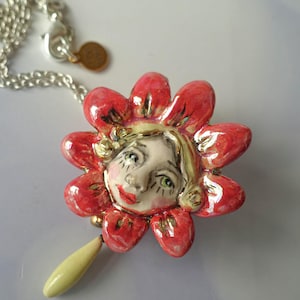 May include: A ceramic pendant necklace featuring a central face surrounded by red petals with gold accents. The face has painted features, including green eyes and red lips. A pale yellow teardrop-shaped bead hangs from the bottom, attached to a silver-toned metal chain with a small circular tag.