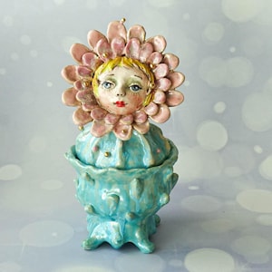 Op de afbeelding: Een keramische pot met deksel met een gebeeldhouwde figuur met een bleek gezicht, blond haar en rode lippen, omringd door een grote roze bloem. De basis is een gestructureerde, lichtblauwe, cactusachtige houder met kleine stekels en glans.