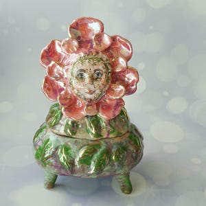 Puede incluir: Un tarro de cerámica con tapa que presenta un rostro esculpido rodeado de grandes pétalos de flores de color rosa iridiscente. La base es un recipiente redondeado de tres patas decorado con hojas verdes y detalles dorados, creando una escultura botánica.