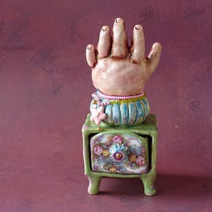 Pode incluir: Uma escultura de cerâmica com uma mão pálida e texturizada sobre uma base de riscas azuis e brancas, adornada com um colar de contas cor-de-rosa e um pendente de flor, sobre uma cómoda miniatura verde-clara com padrões florais.