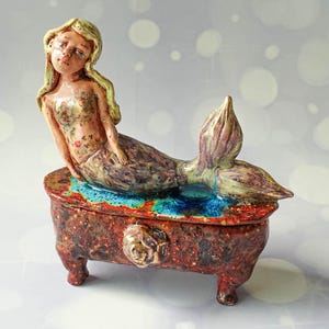 Puede incluir: Un joyero de cerámica con una sirena esculpida de cabello rubio y torso con estampado floral sobre la tapa. La caja es rectangular, tiene cuatro patas, un acabado rojo moteado oscuro y un pomo en forma de rosa en el panel frontal.