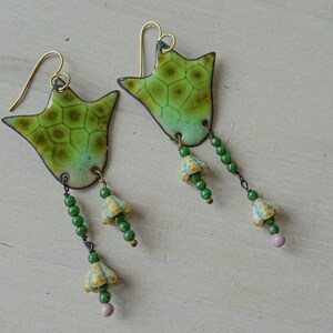Puede incluir: Un par de pendientes hechos a mano con un dise&ntilde;o verde en forma de hoja y un patr&oacute;n texturizado. Los pendientes tienen ganchos dorados y cadenas colgantes con cuentas verdes, peque&ntilde;as flores amarillas y azules y una cuenta rosa al final.