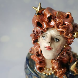 Op de afbeelding: Een keramieken buste van een persoon met krullend roodbruin haar versierd met kleine goudkleurige sterren. Het figuur heeft een lichte huid, blauwe ogen en rode lippen, en draagt een donkerblauw kledingstuk met goudkleurige accenten en een halve maan.