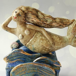 Peut inclure: Une figurine de sirène en céramique avec de longs cheveux flottants, des accents en forme de coquillages et une queue texturée, reposant sur une base de vagues bleues sculptées aux bords dorés. La figure présente des détails peints.
