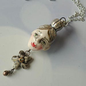 May include: A ceramic pendant necklace featuring a sculpted human head with brown hair, green eyes, and red lips. A small grey five-petaled flower charm with a central amber-coloured bead hangs from the chin, attached to a silver-toned metal chain.