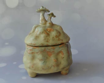 May include: A ceramic jar with a lid shaped like a mushroom. The jar is green and brown with a textured surface. The lid has two mushrooms on top.