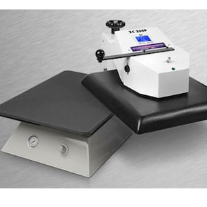 May include: A white heat press with a digital display and a black heat plate. The heat press is on top of a gray base with a black heat plate. The heat press is labeled "JC-205P".