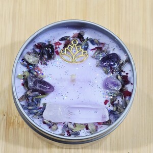 May include: A round, lavender-colored candle in a silver tin. The candle is decorated with amethyst crystals, dried flowers, and a gold-colored lotus charm. A large, clear crystal is embedded in the wax.