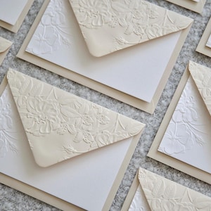 May include: A collection of cream-colored envelopes featuring a triangular flap with a raised, embossed floral pattern. Each envelope is paired with a plain white card insert, showcasing a textured surface and a classic, clean design aesthetic.