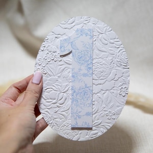 Puede incluir: Una pieza decorativa blanca de forma ovalada con un patrón floral en relieve. Un número uno con estampado floral azul y blanco está adherido al centro. La pieza está sujeta por una mano con uñas pintadas.