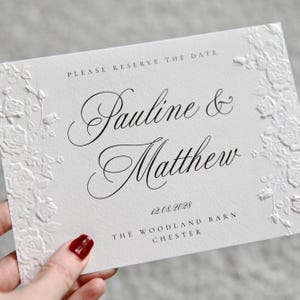 May include: A white rectangular card with embossed floral borders on the left and right sides. The text reads: PLEASE RESERVE THE DATE, Pauline & Matthew, 12.08.2028, THE WOODLAND BARN, CHESTER. The card features black script and serif typography.