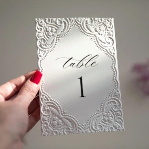 Op de afbeelding: Een witte tafelnummerkaart met een reliëf bloemenrand. Het woord "table" is cursief geschreven en het nummer "1" is gedrukt in een vetgedrukte, zwarte letter. De kaart is rechthoekig.