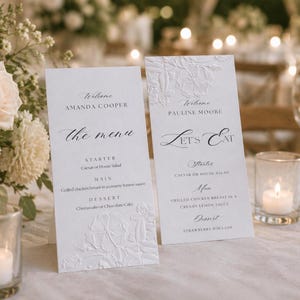 Op de afbeelding: Twee witte trouwmenukaarten met reliëf bloemmotieven. De kaarten hebben elegante kalligrafie met de namen "Amanda Cooper" en "Pauline Moore". Menu-items zijn Caesar salade, gegrilde kip en dessert opties.