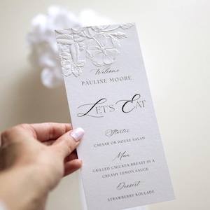 May include: A rectangular white menu card featuring an embossed floral design at the top. The text reads: Welcome, PAULINE MOORE, LET'S EAT, Starter, CAESAR OR HOUSE SALAD, Main, GRILLED CHICKEN BREAST IN A CREAMY LEMON SAUCE, Dessert, STRAWBERRY ROULADE.