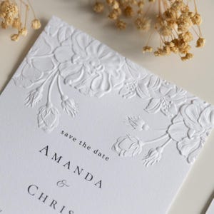 Op de afbeelding: Een witte rechthoekige save the date kaart met een reliëf bloemenpatroon langs de boven- en rechterrand. De tekst luidt save the date gevolgd door de namen AMANDA & CHRIS in een zwart schreeflettertype gecentreerd op het papier.