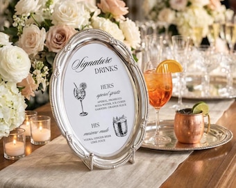 May include: An oval, silver-colored sign with the words "Signature DRINKS" in elegant script. The sign lists two cocktail recipes: "Aperol Spritz" and "Moscow Mule." The sign is displayed on a table with floral arrangements and drinks.