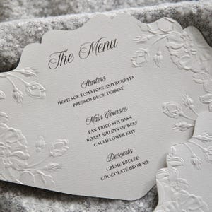 May include: A white scalloped menu card featuring embossed floral patterns along the edges. The text reads The Menu, Starters, Heritage Tomatoes and Burrata, Pressed Duck Terrine, Main Courses, Pan-fried Sea Bass, Roast Sirloin of Beef, Cauliflower Kyiv, Desserts, Crème Brûlée, and Chocolate Brownie.
