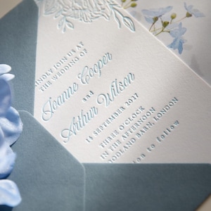 May include: A white wedding invitation card with embossed light blue floral details and blue text reading Joanne Cooper and Arthur Wilson, 14 September 2027, Three O'Clock, Woodland Barn, London, tucked into a dusty blue envelope with a pointed flap.