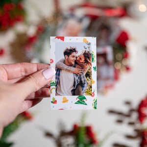 Può includere: Una piccola stampa fotografica rettangolare con una coppia che si abbraccia, con un bordo a tema natalizio di alberi verdi, bacche rosse e campane gialle. La foto è tenuta in mano, con uno sfondo sfocato di decorazioni natalizie.