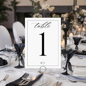 May include: A white table number card with the word "table" in cursive and the number "1" in a bold, black font. The card is held in a silver metal holder. The table is set with glassware, black napkins, and silverware.