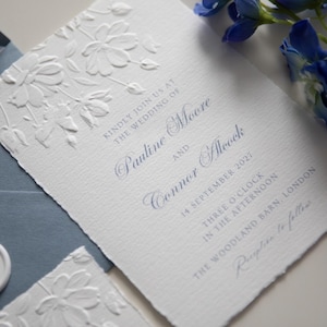 May include: A white wedding invitation card with deckled edges and embossed floral patterns. The text reads: Kindly join us at the wedding of Pauline Moore and Connor Alcock, 14 September 2027, three o'clock in the afternoon, The Woodland Barn, London, Reception to follow.