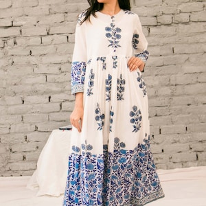 Könnte beinhalten: Ein weißes Maxikleid mit blauem Blumendruck. Das Kleid hat einen Knopfleisten vorne und eine geraffte Taille. Der Rock ist gestuft und der Saum ist ungleichmäßig.