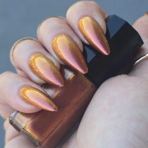 May include: A hand displays long, almond-shaped nails coated in a metallic, colour-shifting nail polish. The polish shifts from gold to pink. A black nail polish bottle is held, matching the nail colour.