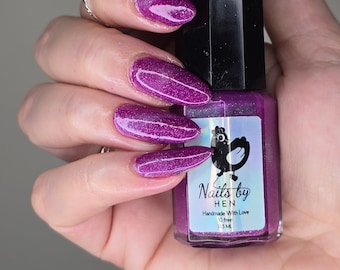 May include: A hand holding a bottle of purple nail polish with a holographic shimmer. The bottle is labeled "Nails by Hen" and says "Handmade with Love 10 free 13.5 ML".