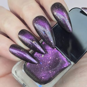 May include: A hand displays long, pointed, purple and black glitter nail polish. The nail polish bottle is square with a black cap and contains matching glitter polish. The nails are a dark purple with a glittery, iridescent finish.