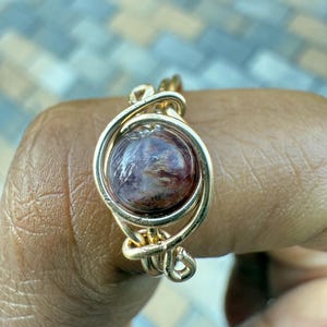 May include: A gold-colored wire-wrapped ring featuring a round, reddish-brown gemstone. The ring's design includes intricate wire detailing around the stone, creating a unique and handcrafted look. The ring is shown on a hand.