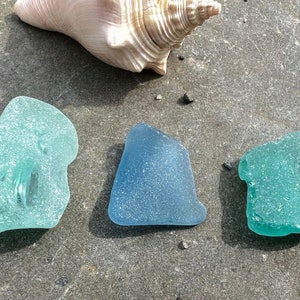 May include: Three pieces of sea glass, one light blue, one dark blue, and one green, on a gray surface.