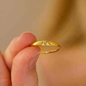 May include: A gold ring with a starburst design is held between fingers. The ring has a smooth, rounded band and a slightly raised, oval-shaped top with the starburst pattern. The ring is held against a neutral background.